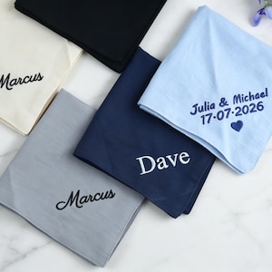 Op de afbeelding: Een verzameling van vijf vierkante stoffen zakdoeken in crème, zwart, lichtblauw, marineblauw en grijs. De crème en grijze doeken zijn voorzien van de geborduurde naam Marcus. De marineblauwe doek heeft de naam Dave. De lichtblauwe doek toont Julia & Michael 17-07-2026 met een hartje.