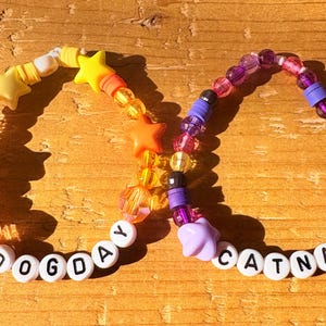 May include: Two beaded bracelets with the words "DOGDAY" and "CATNAP". One bracelet features orange and yellow star beads, while the other has purple and clear beads. The bracelets are on a wooden surface.