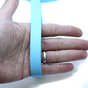 May include: A light blue leather strap, approximately 1 inch wide, is held in a person's hand. The strap is smooth and appears to be made of high-quality leather.