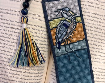 May include: Handmade cross-stitch bookmark featuring a heron design in shades of blue, orange, and yellow. The bookmark has a dark blue border and a tassel with blue, yellow, and orange threads. The bookmark is placed in an open book.