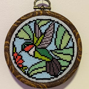 May include: A cross-stitch embroidery of a hummingbird in flight, set within a round wooden hoop. The bird features red, green, blue, and purple hues. The hoop has a decorative wood grain pattern and a metal hanging loop.