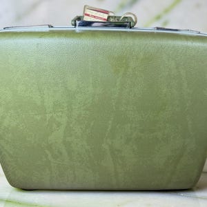 May include: A sage green hard-shell suitcase with a textured finish, featuring a silver-toned metal handle and latch mechanism at the top. A small rectangular tag with illegible text is attached to the handle of the luggage.