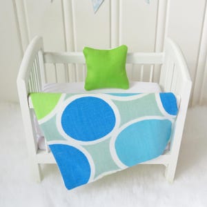 May include: A white miniature crib containing a small lime green square pillow and a blanket featuring a repeating pattern of large blue, light blue, and pale green circles on a white background, draped over the white mattress.