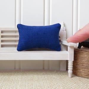 May include: A small, blue cushion sits on a white wooden bench. The cushion is made of a textured fabric and has a rectangular shape.