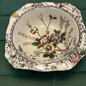 May include: A white ceramic bowl with a floral design. The bowl has a scalloped edge and a brown and white border. The flowers are in shades of pink, purple, yellow, and green.