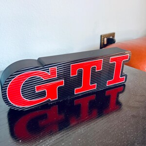 Peut inclure: Un panneau GTI noir et rouge avec un motif en fibre de carbone. Les lettres GTI sont en rouge gras avec un contour blanc. Le panneau est posé sur une surface sombre, reflétant les lettres.