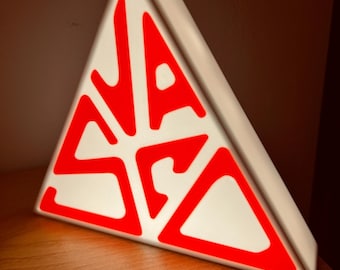 May include: A white triangular sign with red lettering that reads "S.A.S.A." The sign is illuminated from behind.