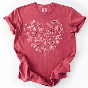 May include: A brick red short-sleeve t-shirt featuring a large heart-shaped graphic on the front. The design consists of white line art illustrations of garden tools, various plants, butterflies, and small yellow and black bumblebees.