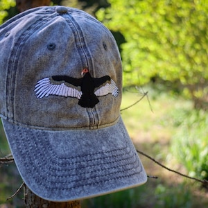 May include: A denim baseball cap with a distressed look. The cap features an embroidered design of a black bird with white wing details and a red head. The hat is set against a blurred green background.