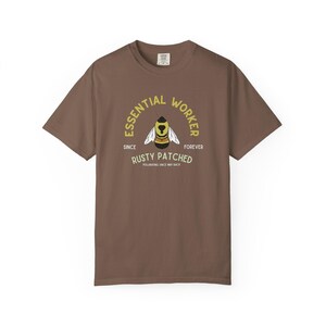 May include: A brown t-shirt with a graphic of a bee wearing a hat, with the text "ESSENTIAL WORKER" above and "RUSTY PATCHED" below. The shirt also has the text "SINCE" and "FOREVER" on either side of the bee.