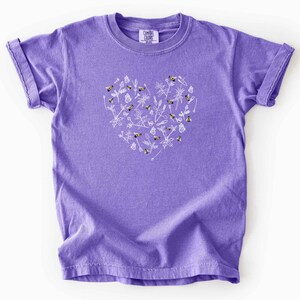 May include: A purple short-sleeve t-shirt featuring a white heart-shaped graphic composed of line drawings of bees, butterflies, and various wildflowers. The shirt has a crew neckline and a Comfort Colors brand label printed on the inner collar.