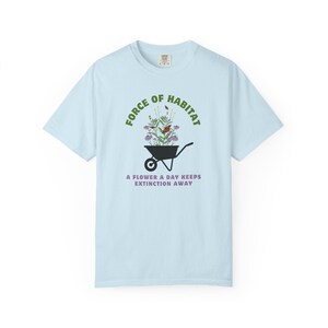 May include: Light blue t-shirt with the text "FORCE OF HABITAT" above a graphic of a wheelbarrow filled with flowers and a butterfly. Below the wheelbarrow is the text "A FLOWER A DAY KEEPS EXTINCTION AWAY".