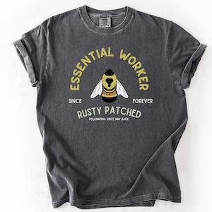Op de afbeelding: Een antracietgrijs T-shirt met korte mouwen en een afbeelding van een bij in het midden. De tekst luidt ESSENTIAL WORKER, SINCE FOREVER, RUSTY PATCHED en POLLINATING SINCE WAY BACK. Het ontwerp gebruikt gele en witte letters op de donkere stof.