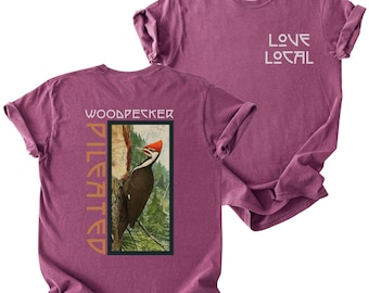 May include: A berry-colored t-shirt featuring a graphic on the back of a pileated woodpecker on a tree trunk with the text "PILEATED WOODPECKER" and a front chest graphic with the text "LOVE LOCAL" in a white, stylized, sans-serif font.