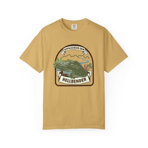 May include: Mustard yellow t-shirt with a graphic featuring a green hellbender salamander, the text "Appalachian Gem" and "Hellbender". The design includes a mountain landscape and is enclosed in a brown border.
