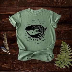 May include: Sage green t-shirt with a black graphic of a hellbender salamander in water, and the word "HELLBENDER" underneath. The shirt has a crew neck and short sleeves, with a slightly faded look. The material is soft and comfortable.