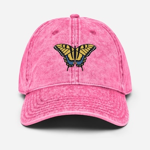 May include: Pink baseball cap with a butterfly design. The butterfly has yellow and blue wings with black outlines. The cap has a curved brim and a fabric strap closure.