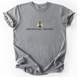 May include: A grey short-sleeve crew neck t-shirt featuring a graphic of a yellow and black bee centered above the black text essential worker. The shirt has a standard fit with a ribbed collar and a visible brand label inside the neckline.