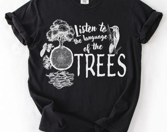 May include: A black short-sleeve t-shirt featuring a white graphic print of a tree, a tree stump cross-section, mushrooms, and a bird. The text reads, Listen to the language of the TREES. The design is centered on the chest of the garment.