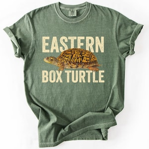 Könnte beinhalten: Ein salbeigrünes Kurzarm-T-Shirt mit einer Grafik einer östlichen Dosenschildkröte mit gelb-braun gemustertem Panzer. Der Text EASTERN BOX TURTLE ist in einer verwaschenen cremefarbenen Schrift über und unter der Schildkröte gedruckt.
