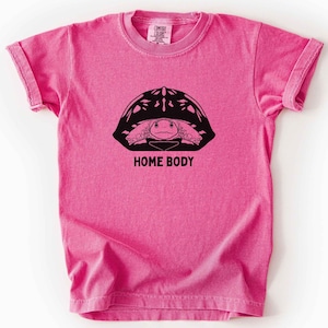 以下が含まれることがあります： 鮮やかなピンク色の半袖Tシャツで、甲羅に入った亀の黒いグラフィックが描かれています。亀の下には黒い大文字でHOME BODYと書かれています。Tシャツはクルーネックで、襟の内側にブランドラベルが見えます。