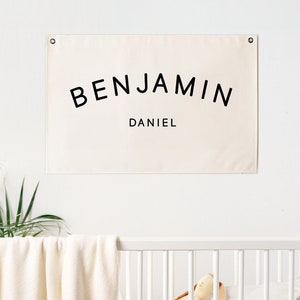 May include: A white canvas banner with black text that reads "BENJAMIN DANIEL" hanging on a white wall above a white crib.