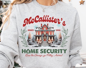 Puede incluir: Una sudadera gris con un diseño navideño que presenta una casa con el texto "McCallister's Since 1990 Home Security Keep the Change, ya Filthy Animal!" impreso en ella.