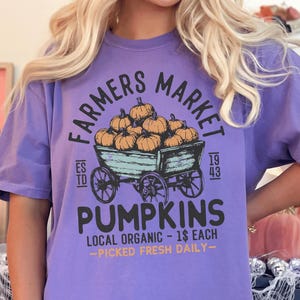 May include: A lilac t-shirt with a graphic design. The design features the text "FARMERS MARKET PUMPKINS" and a wagon filled with pumpkins. The text "LOCAL ORGANIC - PICKED FRESH DAILY" is also present.
