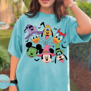 May include: Seafoam green t-shirt with a Halloween-themed graphic of Disney characters. The design includes Mickey and Minnie Mouse, Donald and Daisy Duck, Pluto, and Goofy, all in Halloween costumes. A spider web and bats complete the festive look.