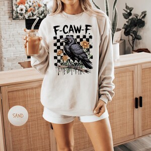 May include: Sand-colored sweatshirt with the text "F-CAW-F" above a black raven perched on a branch, set against a checkered background with yellow flowers. The sweatshirt is a casual, long-sleeved top.