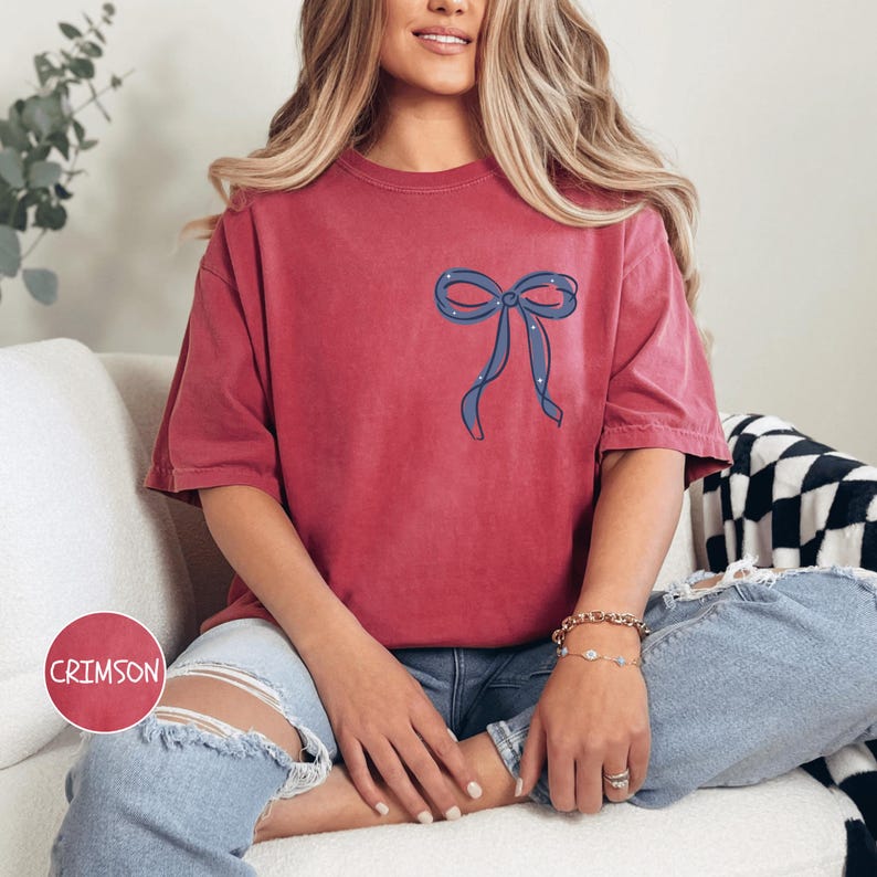 A woman wearing a red t-shirt with a bow design, ripped jeans, and bracelets is sitting on a couch.