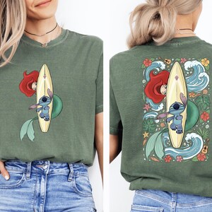 A woman wearing a green t-shirt with a graphic of a mermaid and a surfboard on the back.