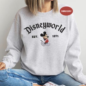 A woman wearing a gray sweatshirt with the text "Disneyworld EST. 1971" and a Mickey Mouse graphic.