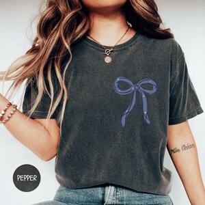 A woman wearing a gray t-shirt with a blue bow design on it, posing against a white background.