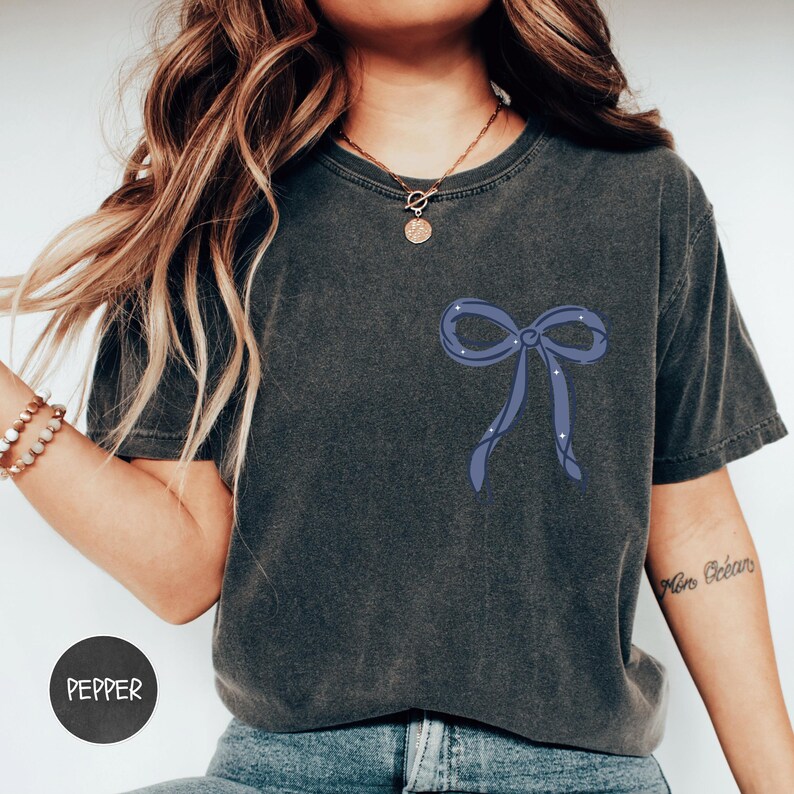 A woman wearing a gray t-shirt with a blue bow design on it, posing against a white background.