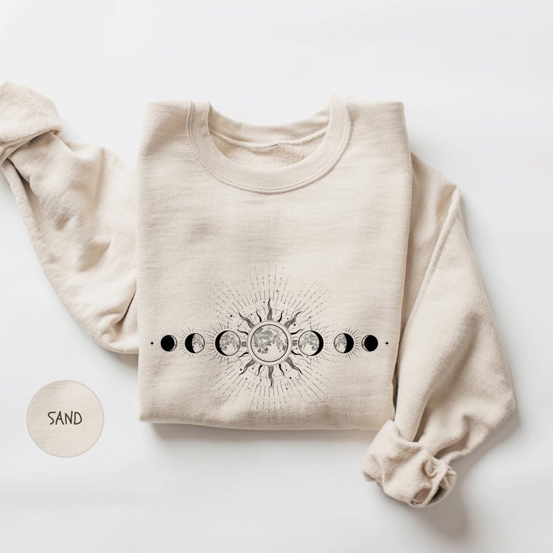 A beige sweater with a graphic design depicting the phases of the moon and sun on the front.