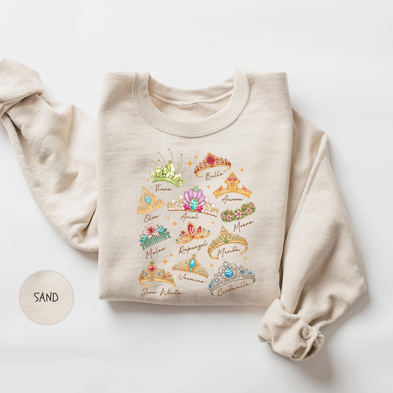 A beige sweater with a colorful graphic design of various crowns and tiaras printed on it.