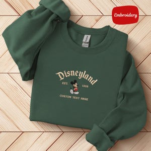 A green sweater with a Disneyland logo and text, laid out on a wooden surface.