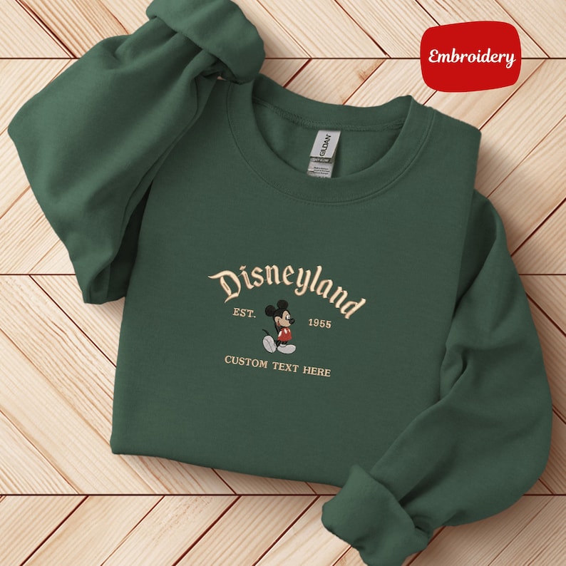 A green sweater with a Disneyland logo and text, laid out on a wooden surface.