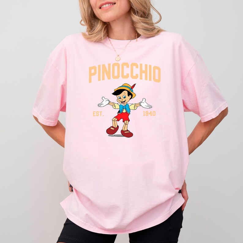May include: A pink t-shirt with a graphic of Pinocchio from the 1940 Disney film. The text "PINOCCHIO" is in yellow above the graphic and "EST. 1940" is below the graphic.