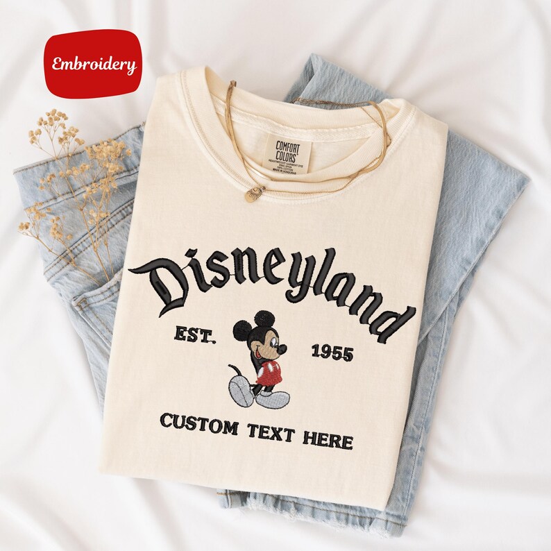 A white t-shirt with a Disneyland logo and text, along with a denim skirt, is displayed on a white background.