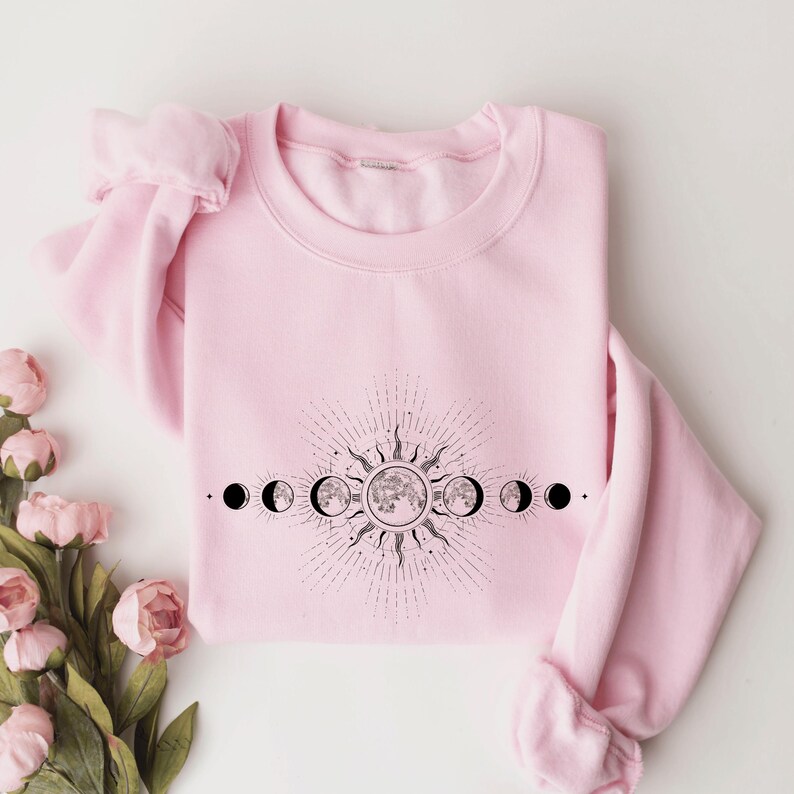 A pink sweatshirt with a graphic design depicting the phases of the moon and sun on it, along with a floral arrangement of pink roses to the side.