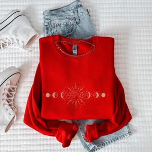 A red sweater with a sun and moon design, a pair of jeans, and white sneakers are neatly arranged on a white surface.