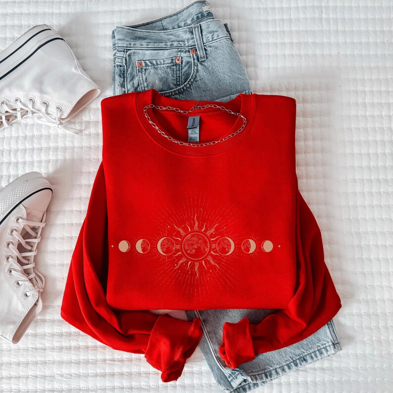A red sweater with a sun and moon design, a pair of jeans, and white sneakers are neatly arranged on a white surface.