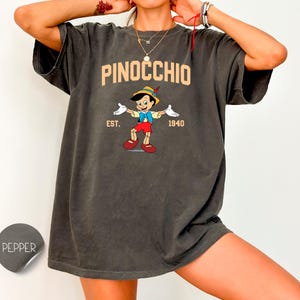 May include: A dark gray t-shirt with a vintage-style graphic of Pinocchio from the 1940 Disney film. The graphic includes the text "PINOCCHIO EST. 1940".