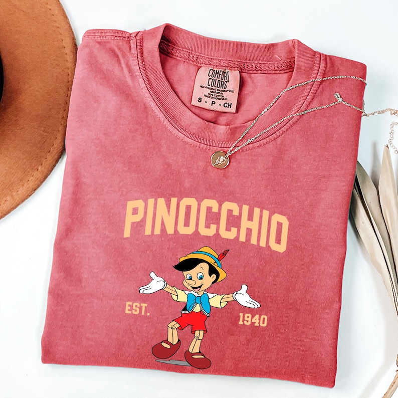 May include: A red t-shirt with a vintage-style graphic of Pinocchio, the Disney character. The text "PINOCCHIO" is in yellow above the image and "EST. 1940" is below the image.