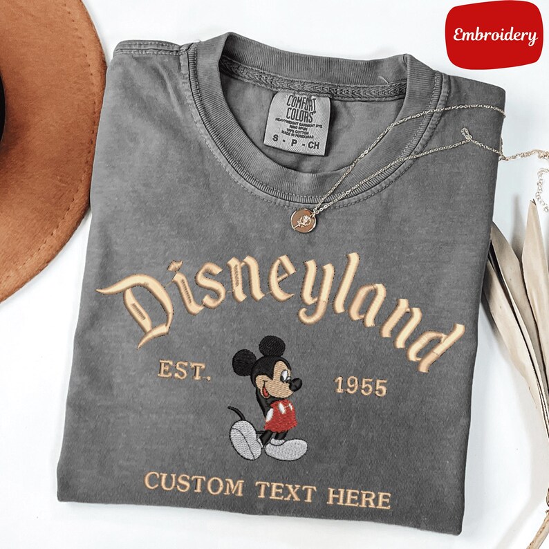 A gray t-shirt with a Mickey Mouse design and text, along with a necklace and a hat, are displayed on a white surface.
