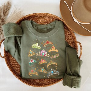 A green t-shirt with a design of various crowns and tiaras is placed in a woven basket, accompanied by a brown hat.