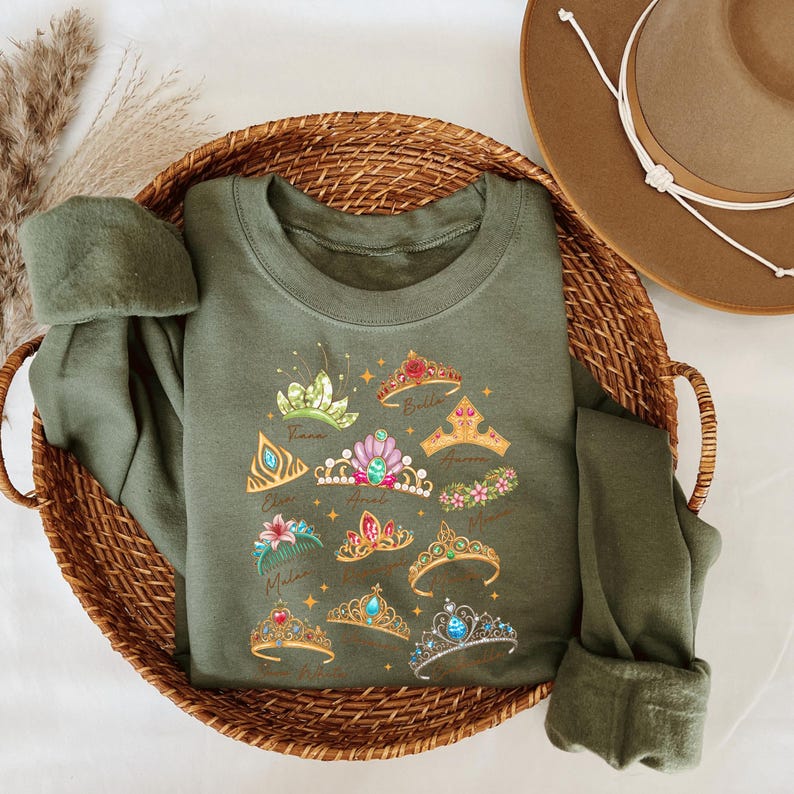 A green t-shirt with a design of various crowns and tiaras is placed in a woven basket, accompanied by a brown hat.