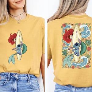 A woman wearing a yellow t-shirt with a graphic of a mermaid and a surfboard on the back.